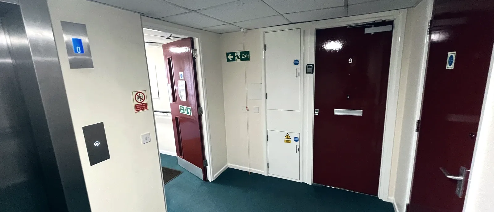 A communal corridor with a maroon fire door, a green running-man fire exit sign, and white service cupboard doors with generic circular safety signage