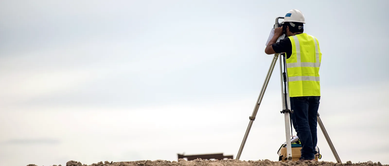 A surveyor using a tripod and total station on a construction site, seen from behind